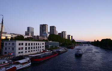 A peaceful riverfront scene showcasing boats alongside modern urban architecture, under a stunning sunset sky.