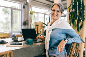 Fototapeta premium Smiling senior freelance woman with gray hair working on laptop at cozy cafe, confident and relaxed