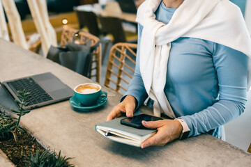Fototapeta premium Close-up of woman holding smartphone and notebook in cafe with laptop and coffee, freelance work
