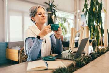 Senior woman with gray hair enjoying peaceful coffee break in cafe with laptop and notebook on table