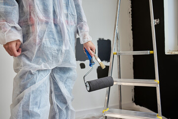 Woman wearing protective coveralls holds black paint roller near painted wall samples and ladder in...