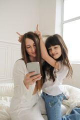 A mother and her young daughter sit close together, holding a smartphone as they chat with someone over video call. Their faces are lit with joy and affection, eyes focused on the screen. 
