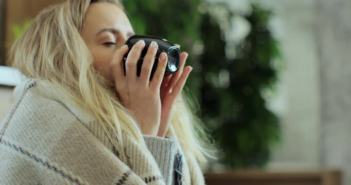 Close up of sick woman sitting at home in blanket with hot tea suffering from fever temperature infection flu headache cold. Young person drinking tea, with seasonal virus symptoms feeling unwell.