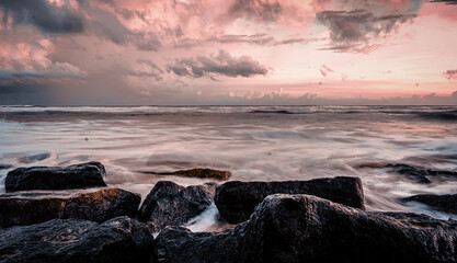 Color long-exposure photograph of ocean waves gently washing over coastal rocks at sunset. A serene, pastel-toned seascape with dramatic clouds and tranquil motion.
