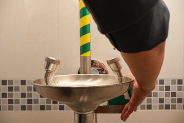A person tests an emergency eye wash station by pressing the activation lever, highlighting safety preparedness procedures in an industrial or laboratory environment with tiled walls.