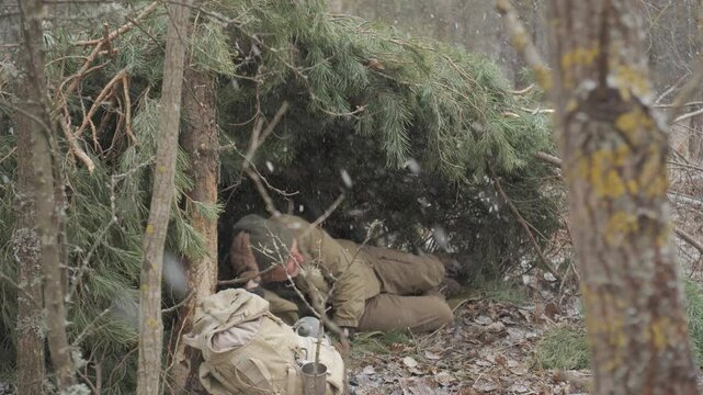 Man sheltering under pine branches during snowfall in forest, Survivalist hides from snow under handmade pine shelter with gear in cold woodland
