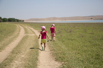 Two young girls in matching pink hats and colorful clothes walk on a grassy path under the sun, one holding a stick.