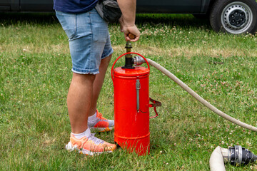 Man operating a red firefighting backpack sprayer (hydronetka) on the grass. The device is connected to a pressure hose and used during fire and rescue training.