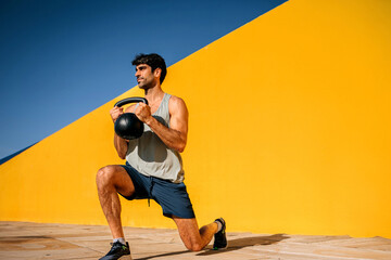 Hispanic athlete performs lunges with kettlebell against vibrant yellow backdrop at outdoor...