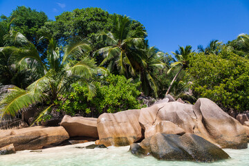 A coast with giant granite stones, trees and palms, the Indian Ocean and blue sky with clouds. La Digue, the Seychelles