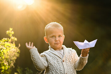 Little boy playing alone in the forest with a white paper boat. Peaceful childhood moment filled...