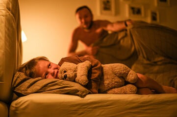Father gently tucks his little child into bed with a teddy bear. Cozy bedtime scene filled with...