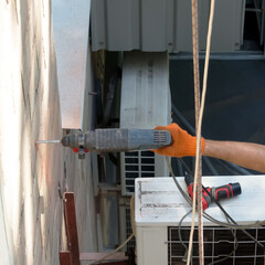 A worker drills a hole in the wall of a house.Red rock dust flies everywhere.Next to the air conditioner lies a spare drill.Using spare tools in construction is a necessary element of the work process