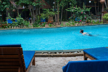 A man is leisurely swimming in a large swimming pool, surrounded by chairs