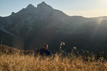 Man resting in tall grass with two dogs - german and australian shepherds - at sunset, with Komovi mountains in background. Autumn colors and golden light create a serene and peaceful atmosphere.
