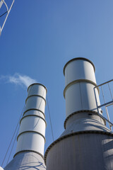 Two tall gray industrial chimneys rise against a clear blue sky. One gently releases a small puff of white smoke. Scene highlights industry and the importance of environmental considerations.