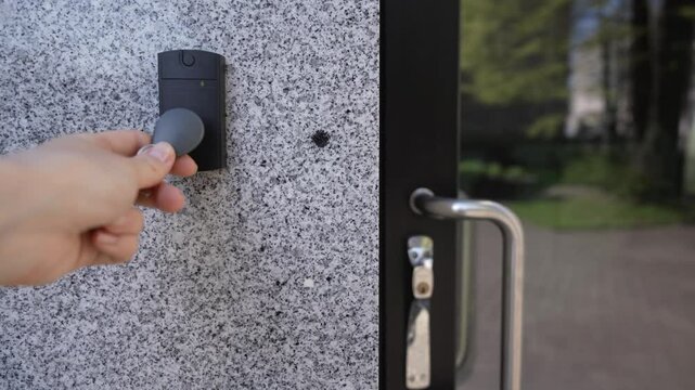 Modern access control concept. Close-up of female hand using key fob on smart door entry device. Red light changes to green. Glass door, granite wall background. Office security in action