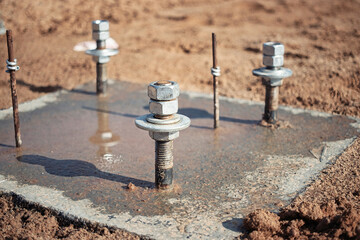 Construction site shows bolts embedded in concrete with water pooling around them during bright daylight