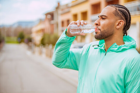 Afro-american athlete with cornrows refreshing drinking a bottle of water after training outdoors.