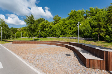 Obraz premium In a pleasant park, a sturdy wooden bench can be found situated right alongside the side of a road, providing a resting spot for visitors enjoying their surroundings and natures beauty