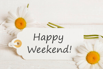 Happy Weekend message on white paper tag with daisies, minimal floral layout on light wood, clean summer concept for digital greeting cards, lifestyle blogs, printable templates, morning wishes