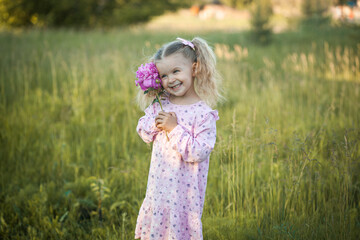 cheerful 3-year-old girl with pigtails in a pink dress with a peony walks in a village meadow