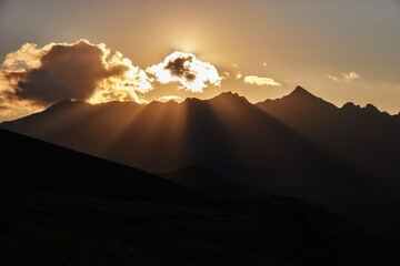 Sunlight breaks through clouds, casting rays over rugged mountain peaks during sunset, creating a...
