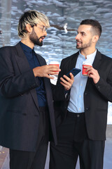 Vertical composition showing two young businessmen in suits having coffee and talking while checking a phone. Ideal for themes of communication, business interaction, and modern workplace. 