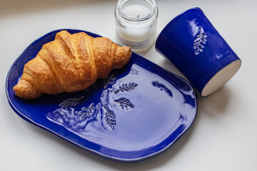 Handmade blue clay plate with croissant on it. Vase with wheat and candles.
