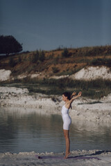 Woman in white sportswear sitting on yoga mat by lake holding foot in stretching pose. Outdoor fitness and flexibility training concept. Design for poster, banner, greeting card, invitation. Side view