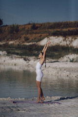 Woman in white sportswear standing on yoga mat by lake stretching arms. Outdoor fitness and healthy lifestyle concept. Design for poster, banner, greeting card, invitation. Side view