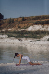 Woman in white sportswear kneeling on yoga mat by lake in stretching position. Outdoor fitness and healthy lifestyle concept. Design for poster, banner, greeting card, invitation. Side view