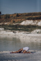 Woman in white sportswear doing child’s pose on yoga mat by lake. Outdoor yoga and relaxation practice. Design for poster, banner, greeting card, invitation. Side view