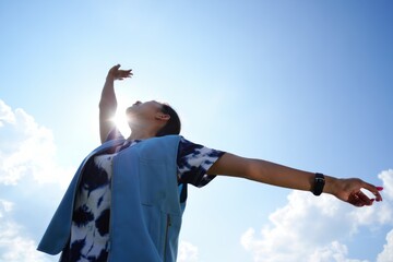 Person stretching arms wide open under clear blue sky with sun shining brightly behind and a few fluffy clouds