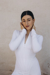 Woman in white zip-up bodysuit touching her face. Studio fashion portrait on beige textured background. Beauty and style concept. Design for poster, banner, greeting card, invitation. Front view