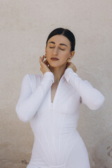 Woman in white zip-up bodysuit with closed eyes touching neck. Studio fashion portrait on beige textured background. Beauty and style concept. Design for poster, banner, greeting card, invitation. 