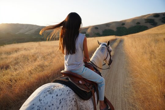 Young woman with long hair riding a white horse with dark spots along a dirt path through a golden dry grass field at sunset in a hilly landscape