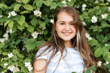 Smiling teenage girl 13 -14 year old with long healthy blonde hair over blooming jasmine bush with white flowers in garden outdoors. Happiness.