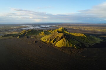 Aerial view of a volcanic mountain covered with green vegetation casting shadows on surrounding dark volcanic desert under a clear sky with scattered clouds