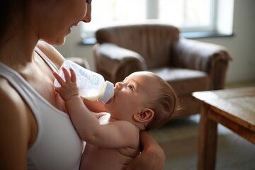 Happy mother holding and feeding her baby with a bottle in a cozy living room filled with natural light