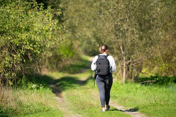 A woman hiking in a verdant forest, wearing dark pants, a light colored long sleeve shirt, and a dark pack, walking away on a path.