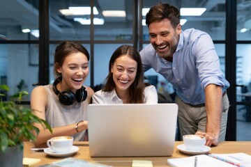 Three cheerful coworkers enjoying a moment of success while looking at a laptop in a modern office setting with coffee cups on the table