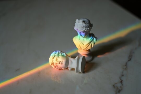 Two classical-style white bust sculptures on a smooth surface illuminated by a vivid rainbow light beam casting colorful reflections and soft shadows