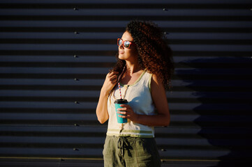 woman with coffee cup enjoys sunshine outdoors wearing sunglasses and casual outfit. sunlight and shadows create dramatic contrast on textured background inspiring summer vibes