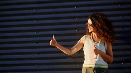 woman giving thumbs up outside. confident woman in casual attire and sunglasses gestures approval against industrial background. bright sunlight emphasizes carefree and positive vibe