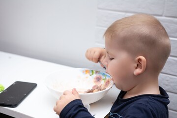A young child eating from a bowl with a spoon, focused and determined, sitting at a white table in...
