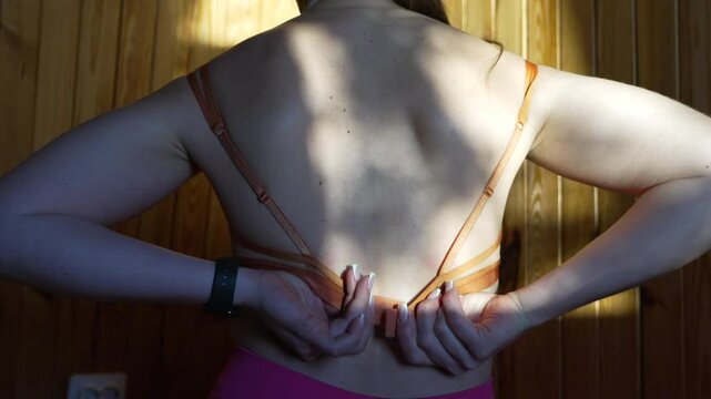 Unbuttoning Bra Back woman unhooks lingerie indoors against wooden wall