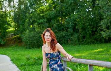Fototapeta premium Woman in blue dress leaning on bridge railing in park. Relaxation and rest concept as woman enjoying her time outdoors. Lush green surroundings. Girl has beautiful body.