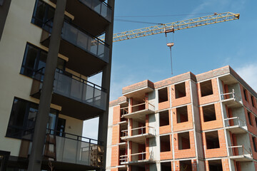 Modern apartment building with dark glass balconies stands next to a new brick structure under construction, highlighted by a tall yellow crane against a clear blue sky.