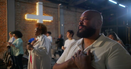 Man standing up from seat in church, placing hands on chest, closing eyes in prayer, illuminated cross in background, community worship and spiritual reflection